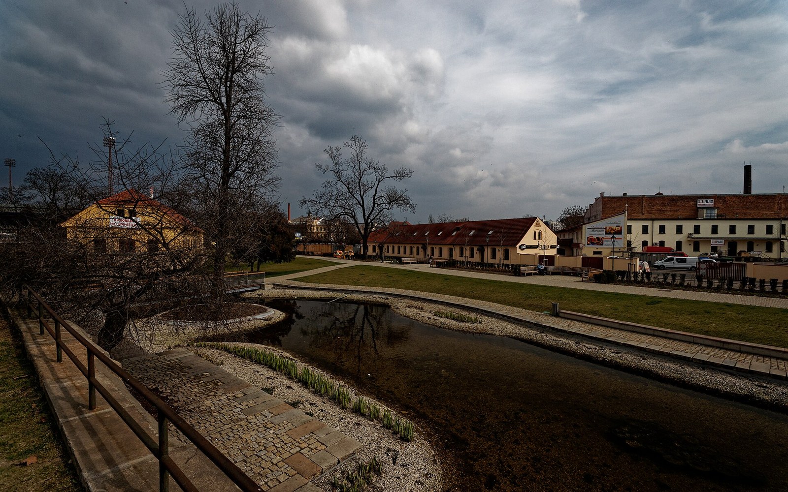 Bare trees line a canal beside old brewery buildings under a stormy sky