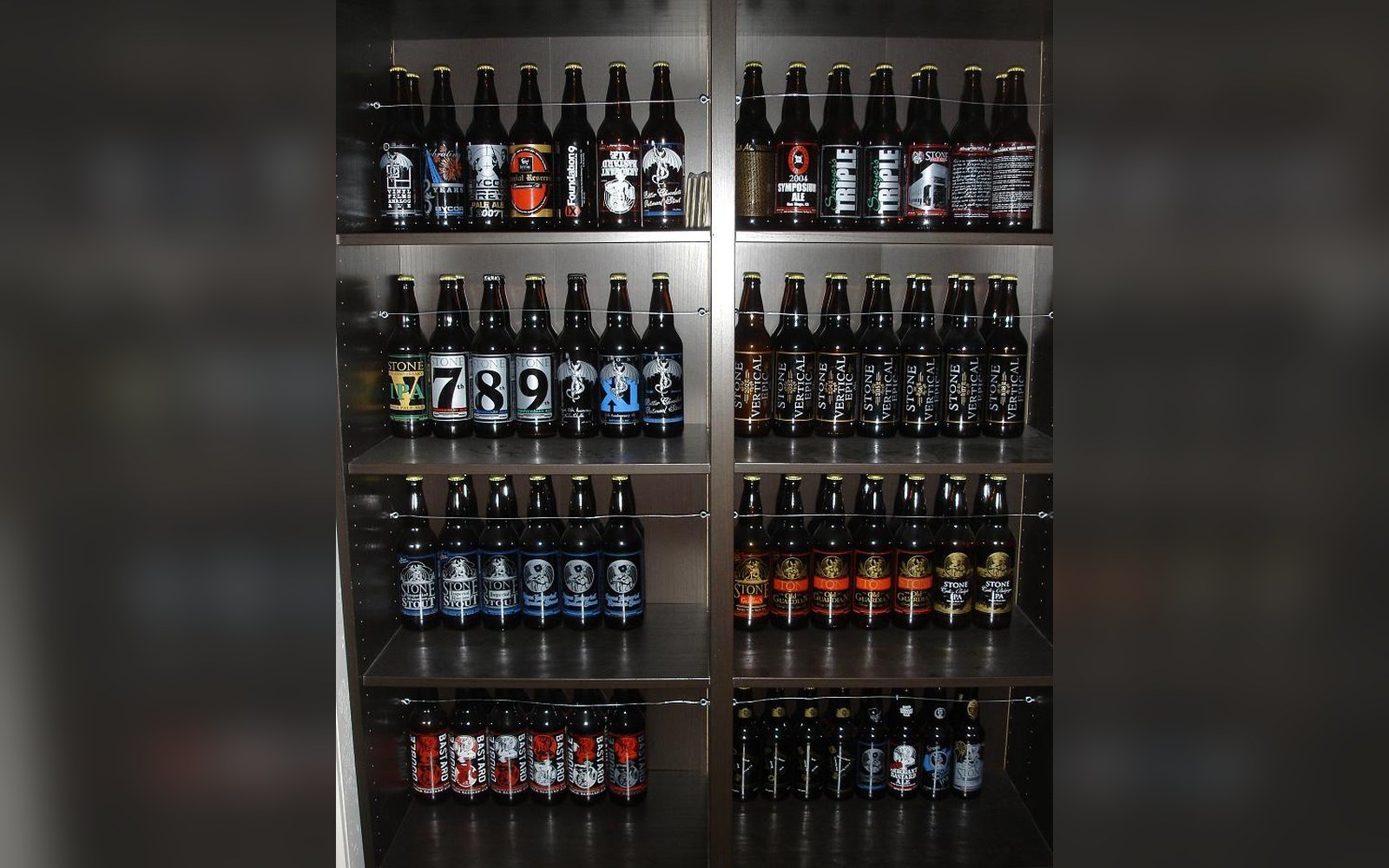 Rows of labeled beer bottles lined up on dark wooden shelves in a cabinet