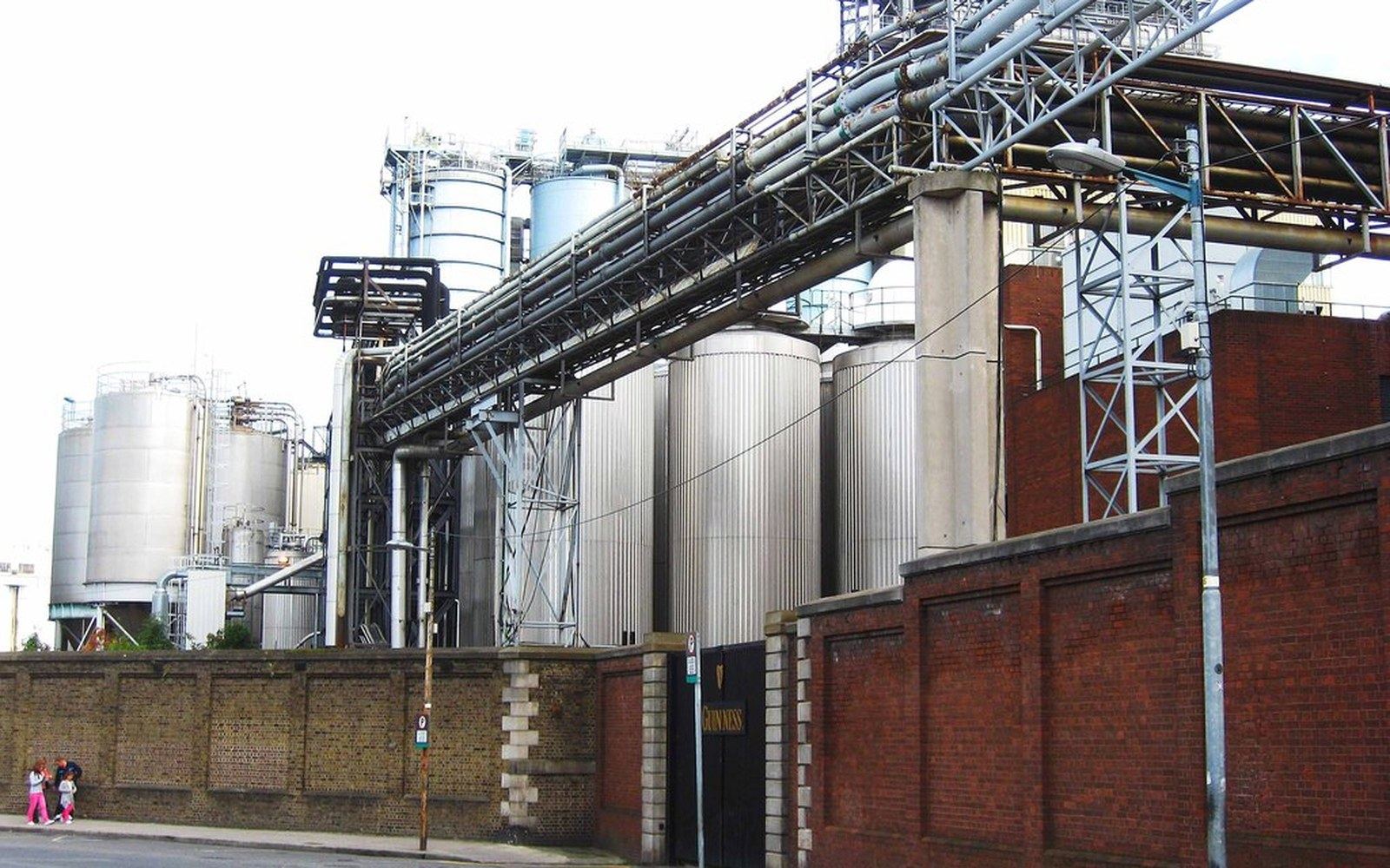 Metal storage silos and elevated pipework rise above a brick perimeter wall at a brewery